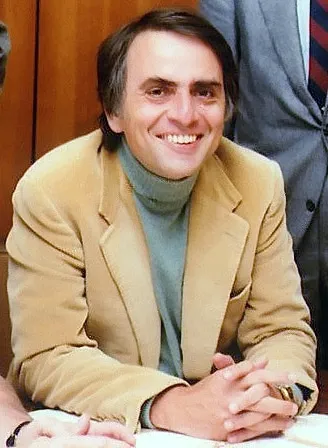 Carl Sagan seated in a tan jacket, smiling with hands folded.