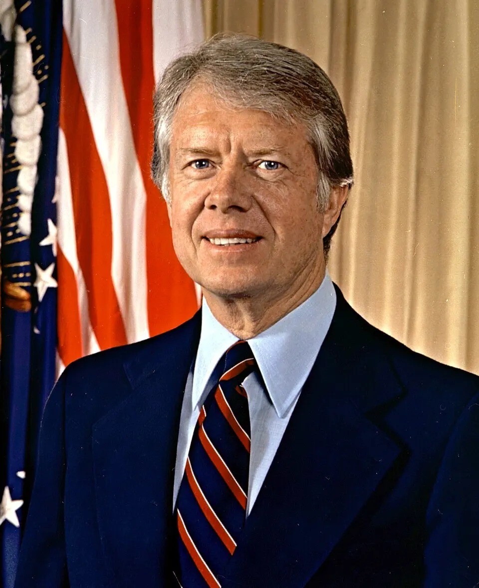 Official 1977 presidential portrait of Jimmy Carter, smiling in a navy suit in front of an American flag.
