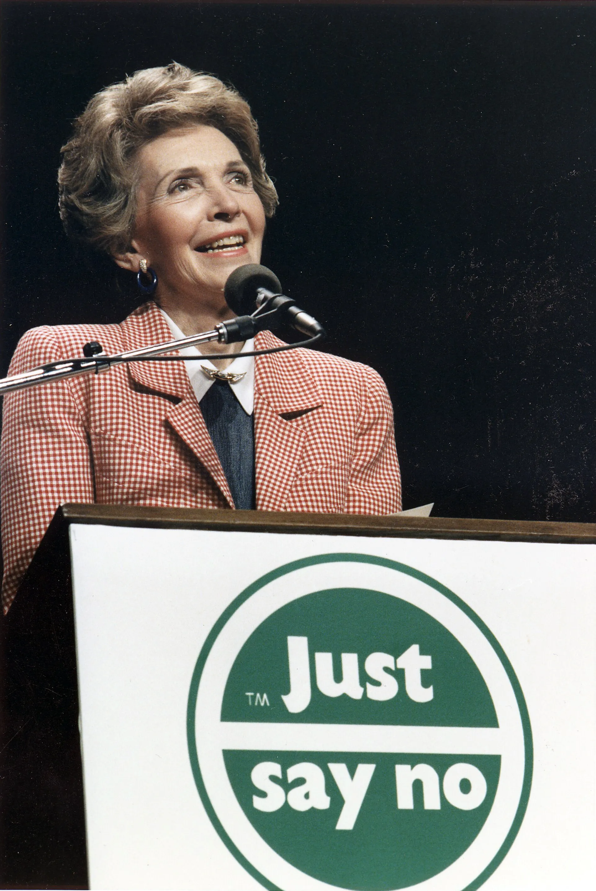Nancy Reagan smiles at a microphone behind a podium bearing a large green “Just say no” logo at a Los Angeles rally in 1987.