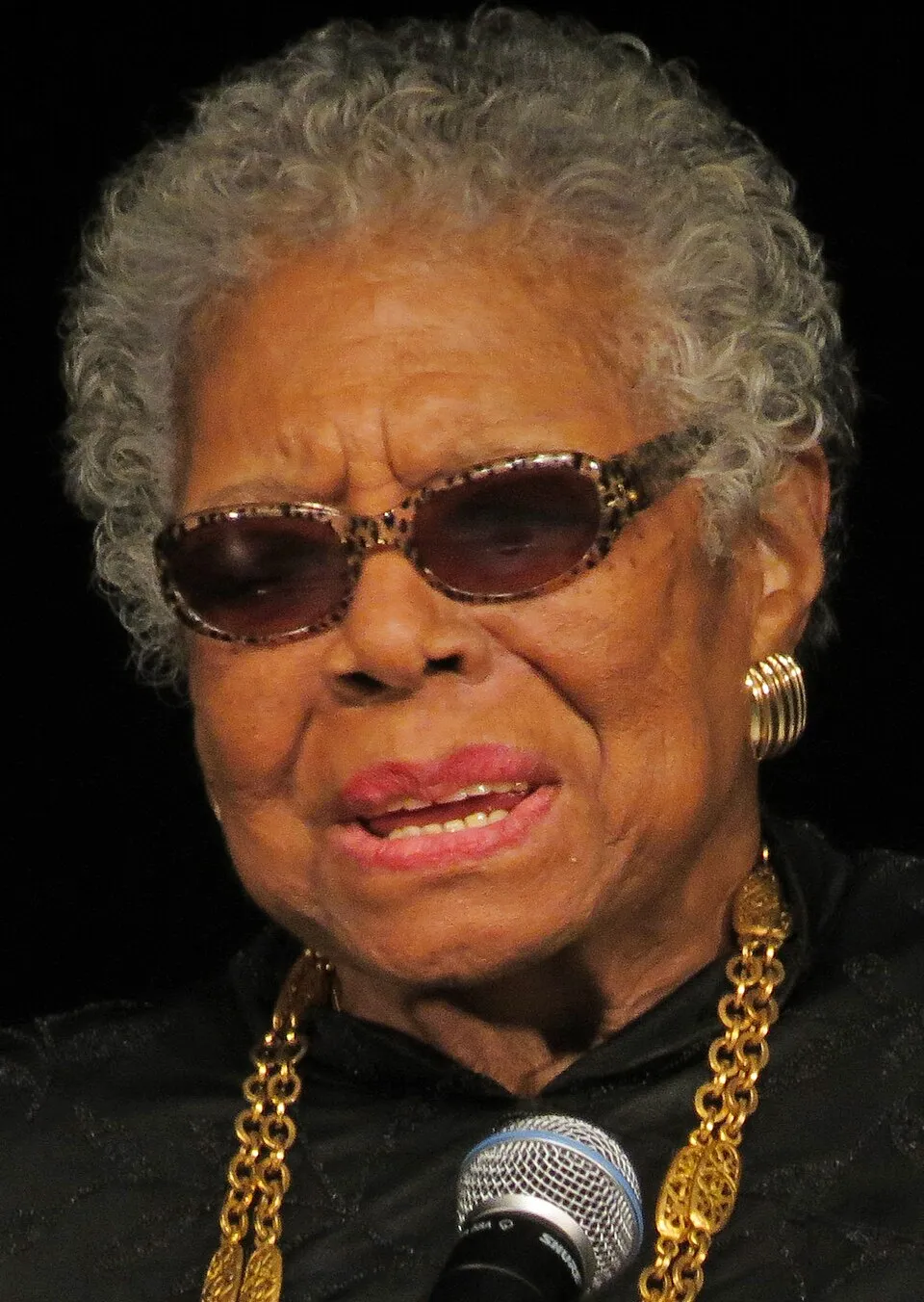 Maya Angelou speaking at a microphone, wearing gold necklaces and tinted glasses.